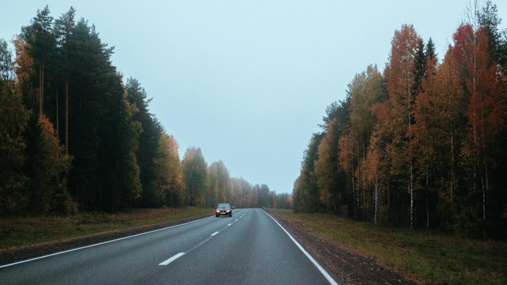 A serene autumn scene of a car driving on a highway flanked by colorful trees.